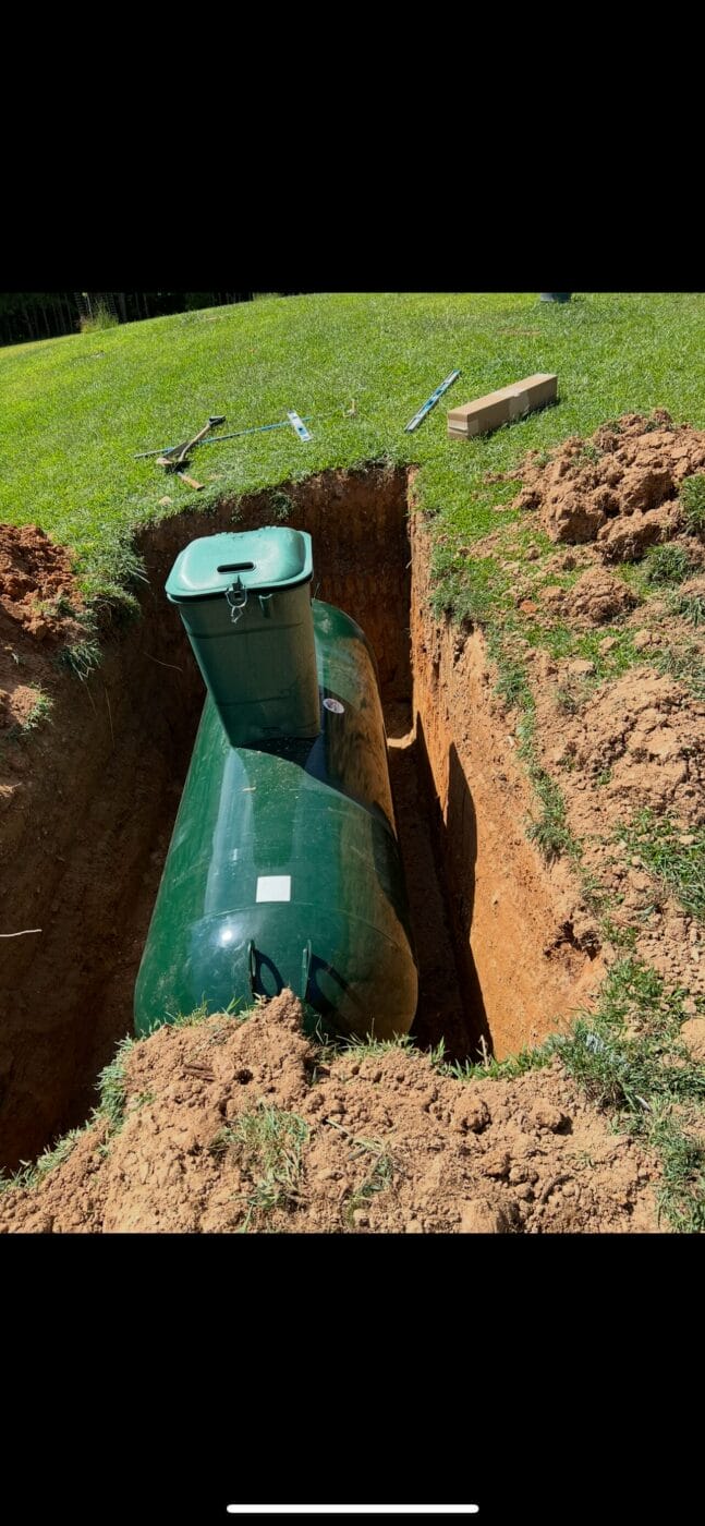 A large green propane tank partially buried in a freshly dug trench in a grassy yard, with tools and a cardboard box nearby.