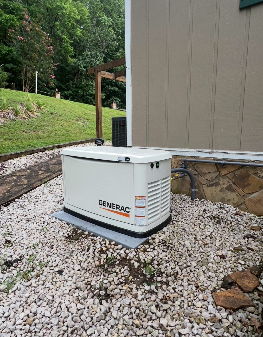 A Generac standby electric generator is installed on a gravel pad next to a house with tan siding and stone trim, with a grassy yard and trees in the background.
