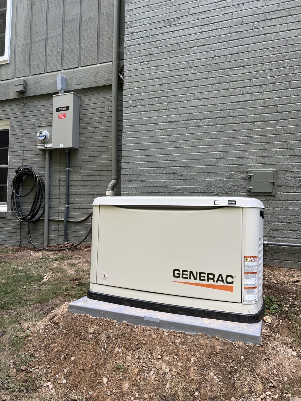 A Generac electric generator is installed on a concrete pad next to a brick house, with electrical wires and a utility box mounted on the wall.