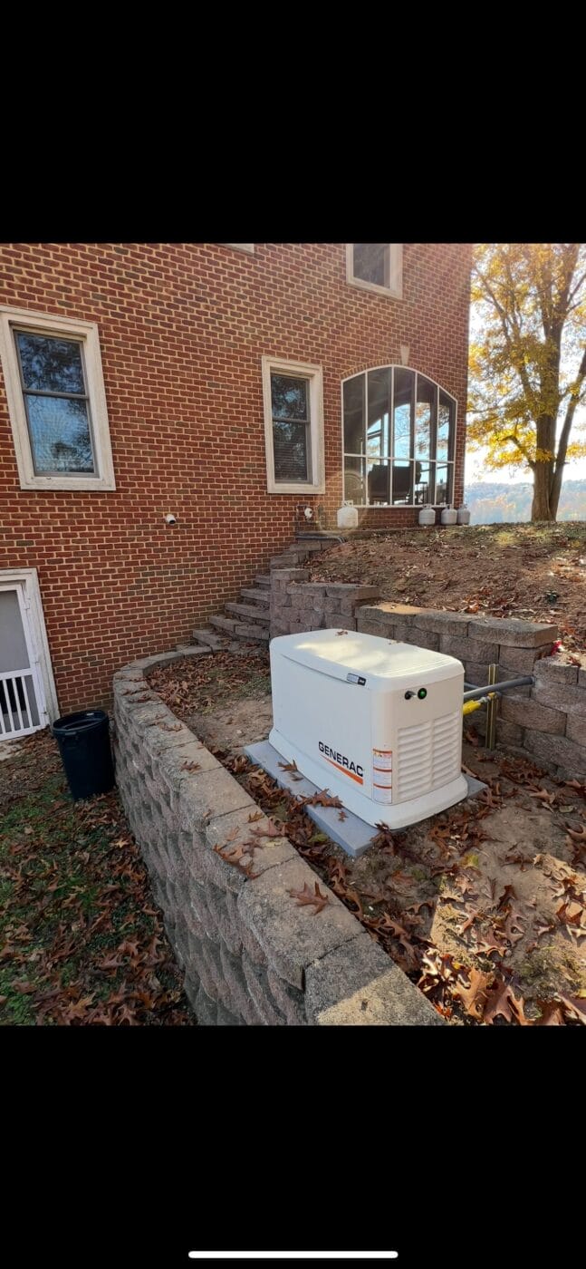 A Generac standby generator is installed next to a brick house, set on a concrete pad within a walled, landscaped area.